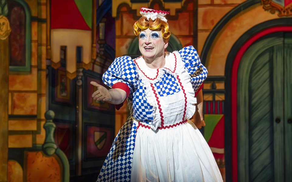 Nanny Nutkins smiles at the audience, she is wearing a blue and white checkered dress and an apron