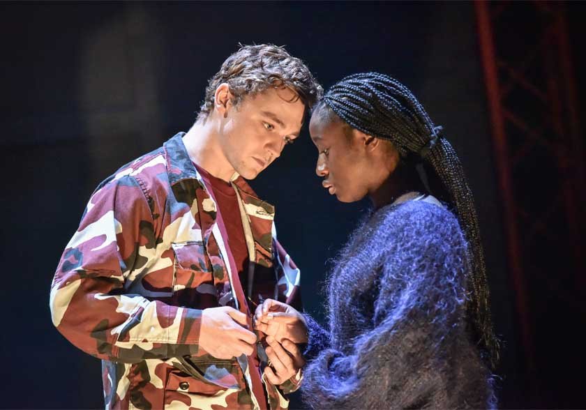A white man in a camo top looks intensely at a black woman in a purple jumper, they hold hands