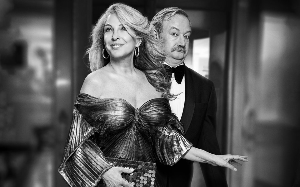 A glamorous man and woman stand together in a posh evening gown and a suit, the woman is smiling and the man looks suspicious