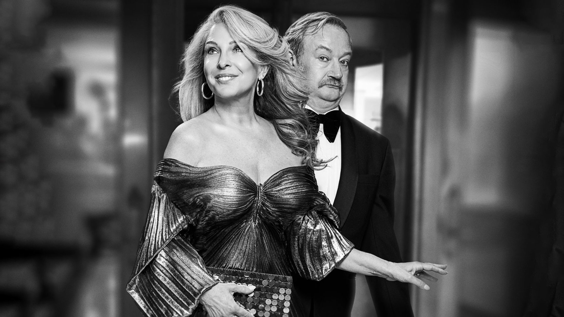 A glamorous man and woman stand together in a posh evening gown and a suit, the woman is smiling and the man looks suspicious