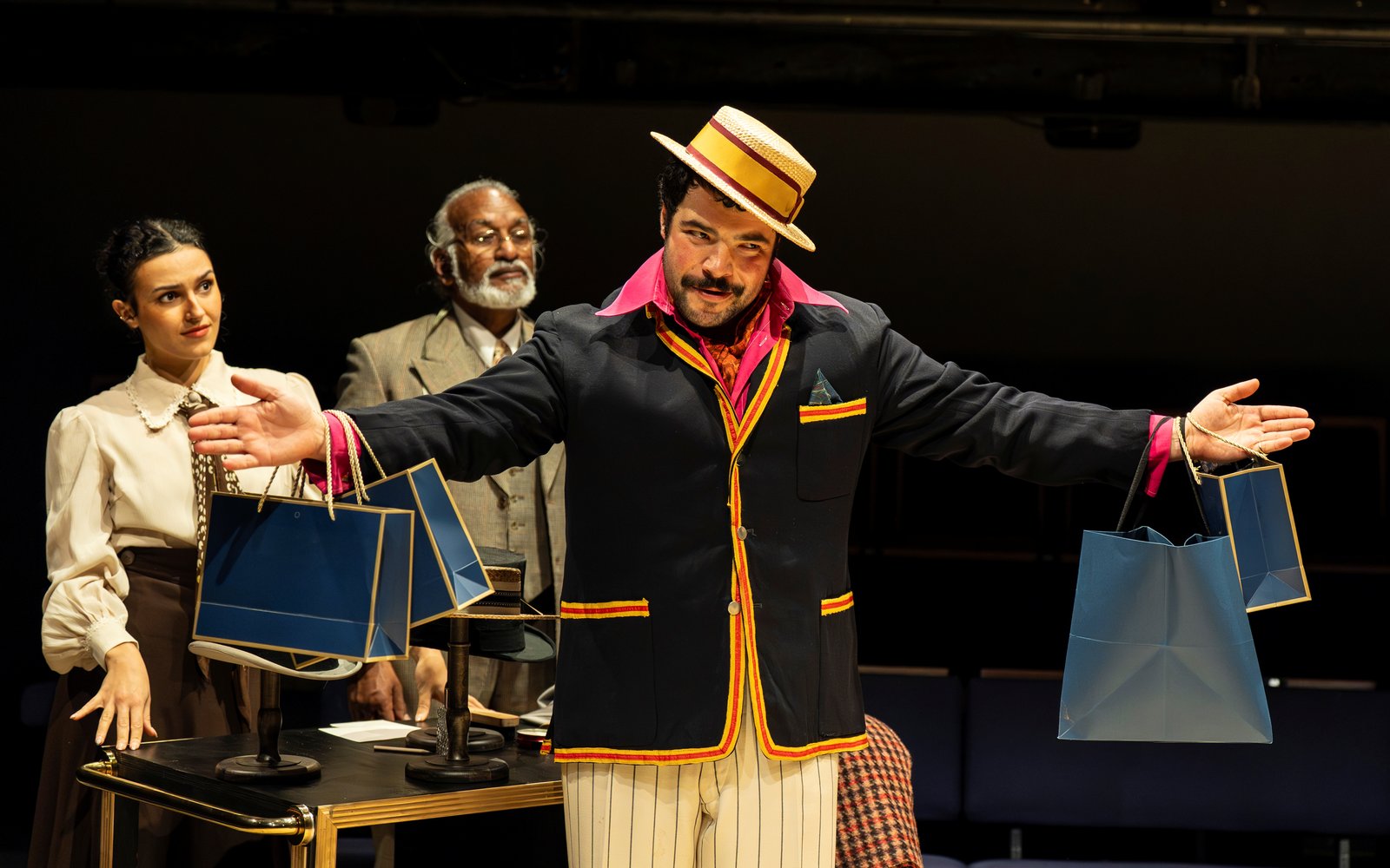 A man in a dark jacket, pink shirt, cream striped trousers and a boater hat smiles mischievously. His arms are outstretched, with two shopping bags in either hand.