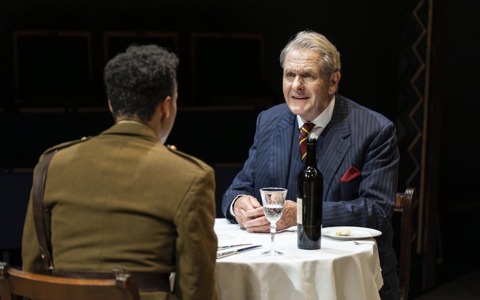 Robert Bathurst, wearing a navy pinstripe suit, sits at a table with a man in military uniform.