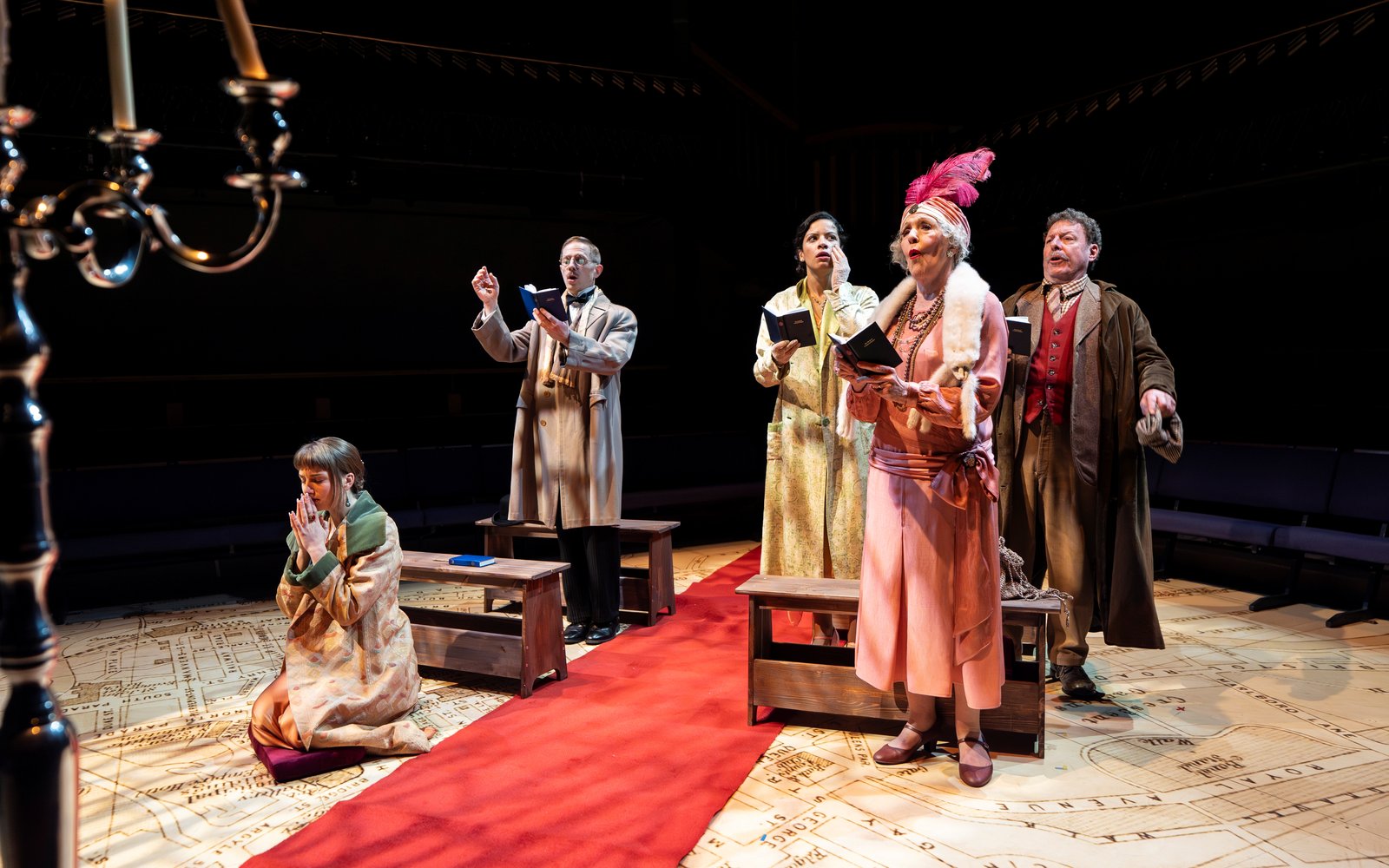 The cast of The Rivals stand in front of pews on either side of the aisle. One woman kneels at the front, praying, while the rest of the cast sing from hymn books.