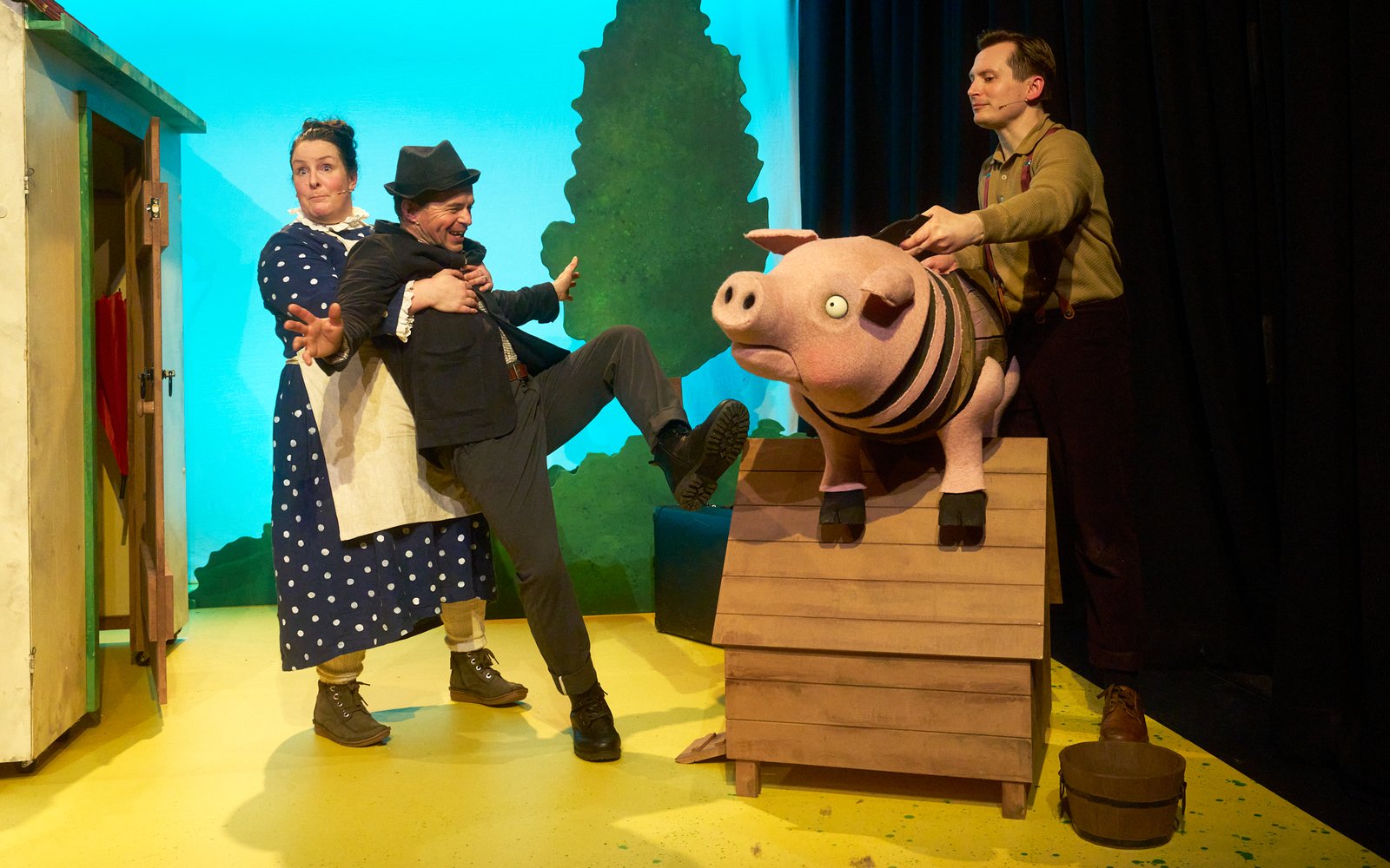 A woman holds a man up whilst he looks shocked and happy as a large puppet pig looks at them