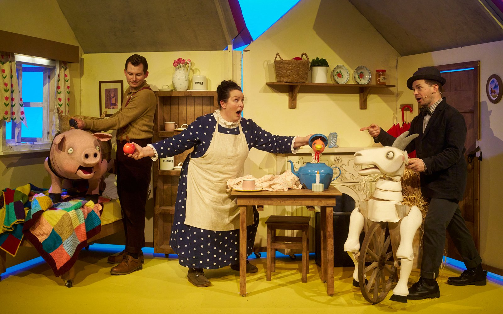 A woman in a polka dot dress and apron in a little cottage, there are puppets of a pig and a goat either side of her