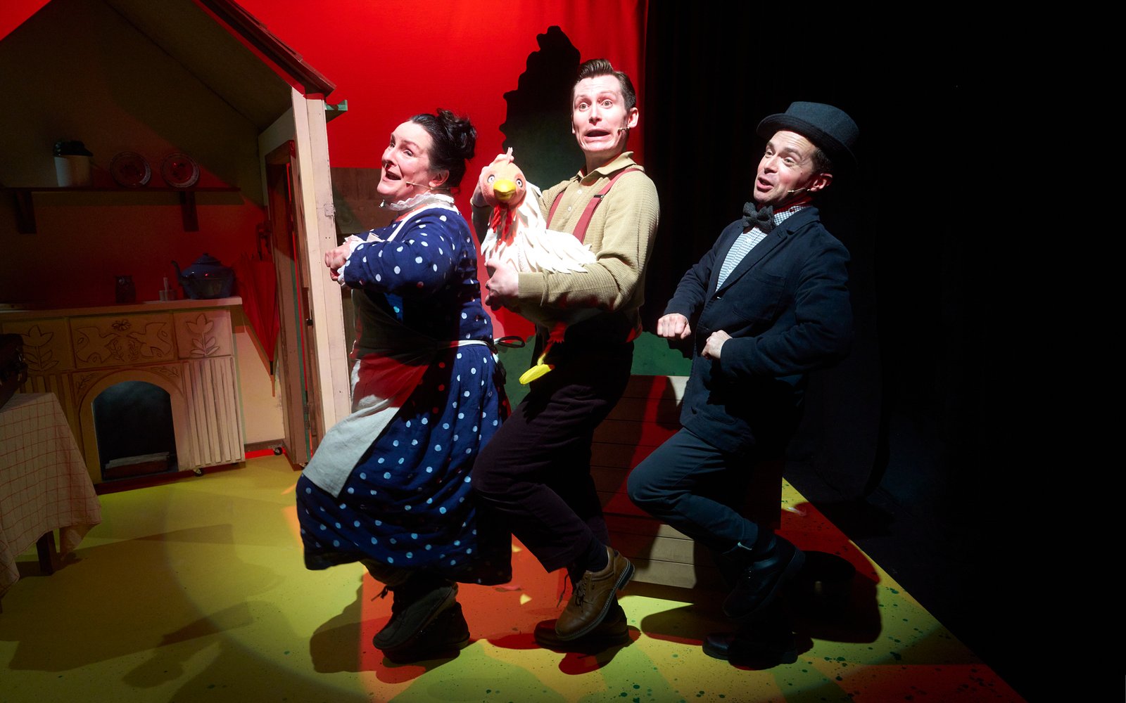 A woman and two men dance in a line, the man in the middle is holding a puppet chicken
