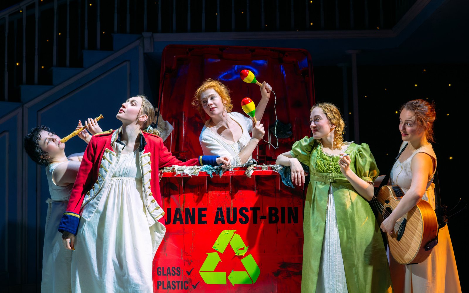 A group of women sing around a large industrial bin, they are shaking maracas and playing the flute