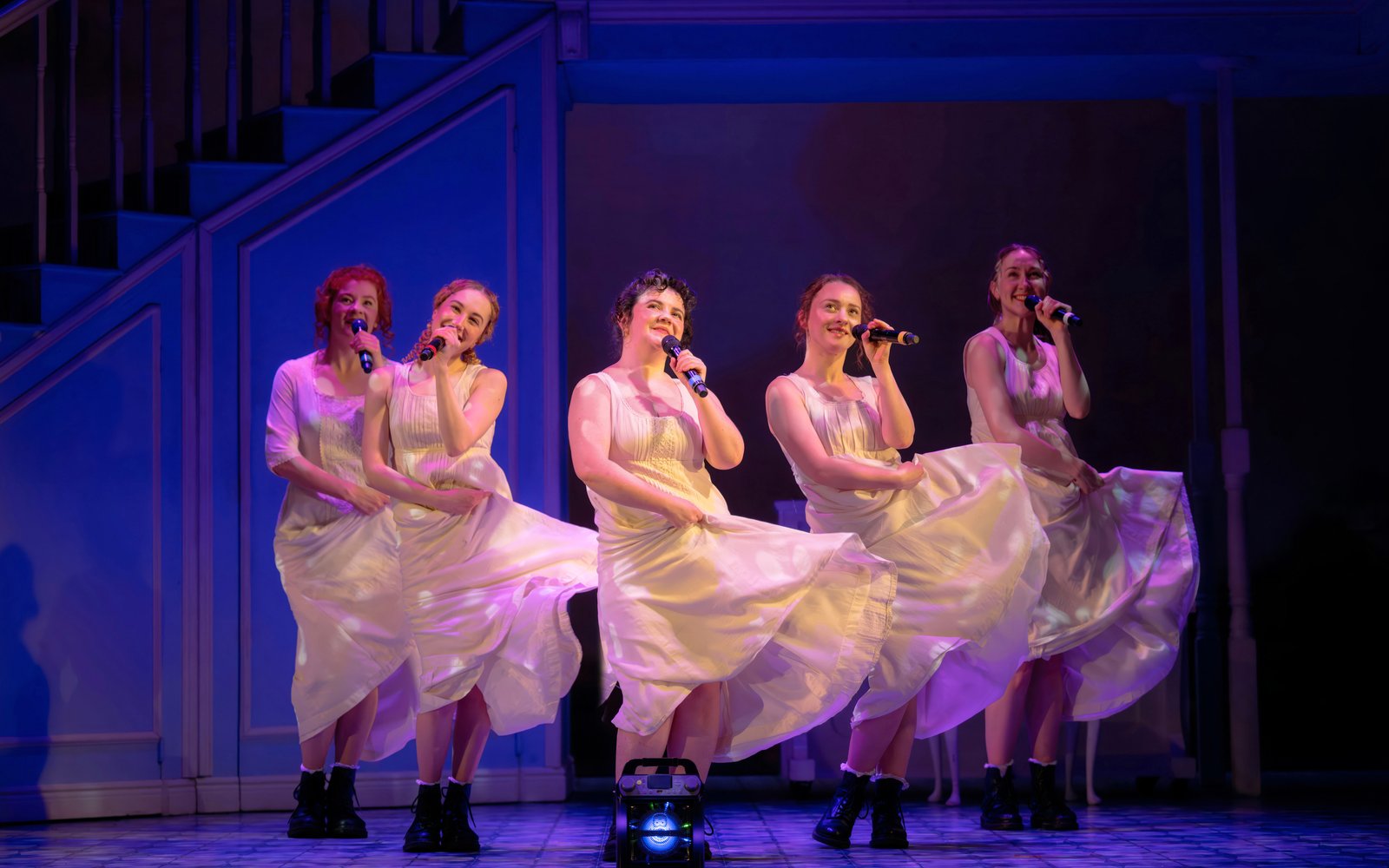 Five women in white dresses sing in unison holding microphones