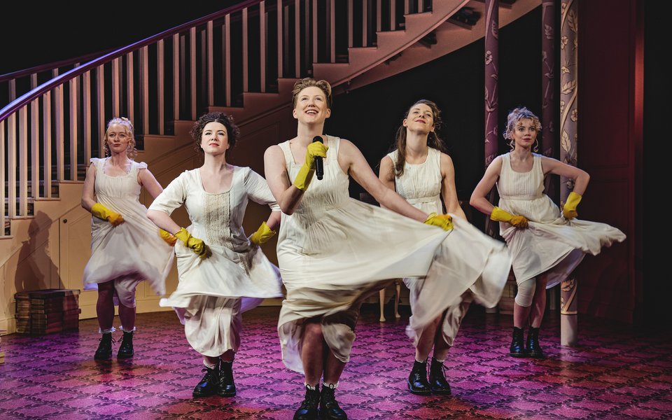 Five women standing in a row, the are wearing white dresses and yellow rubber gloves. One of them is singing into a microphone