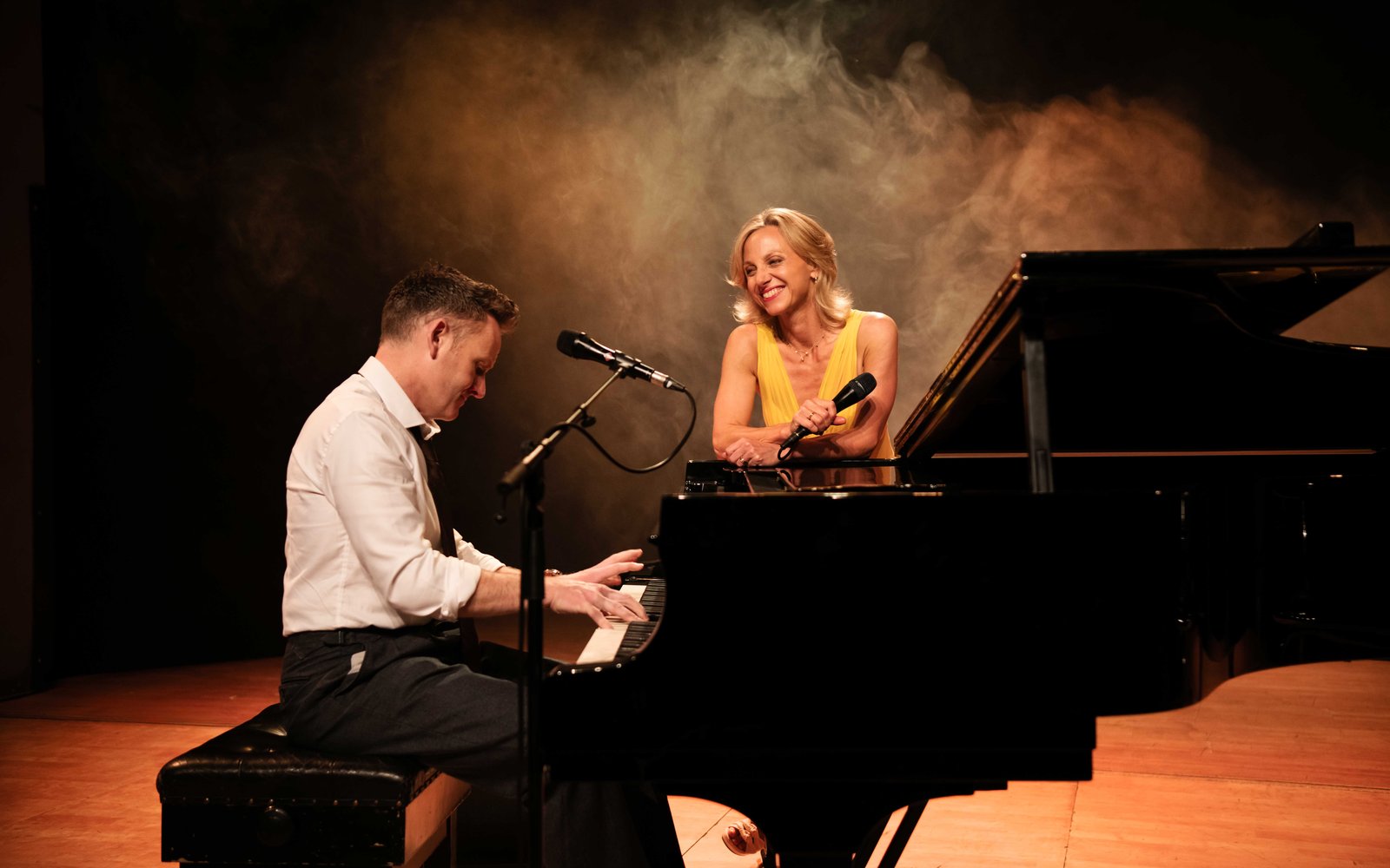 A man sitting at a grand piano playing music, a woman in a yellow dress leans against the piano holding a microphone