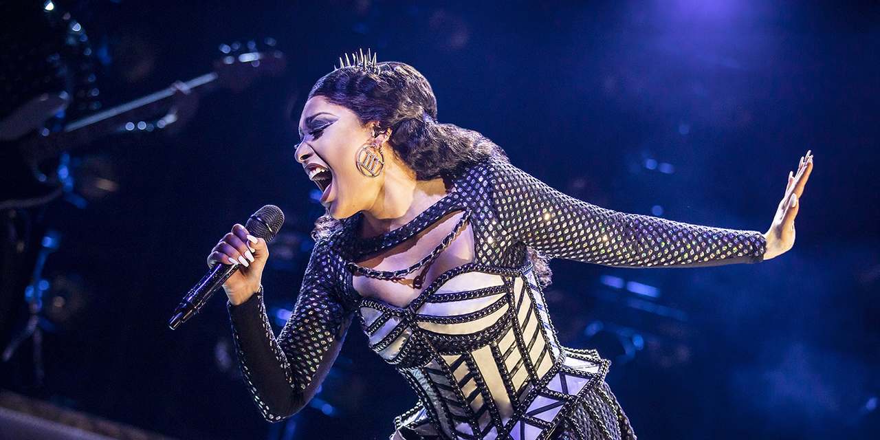 A woman singing passionately into a microphone, she is wearing a spiked crown, big earrings and silver studded dress