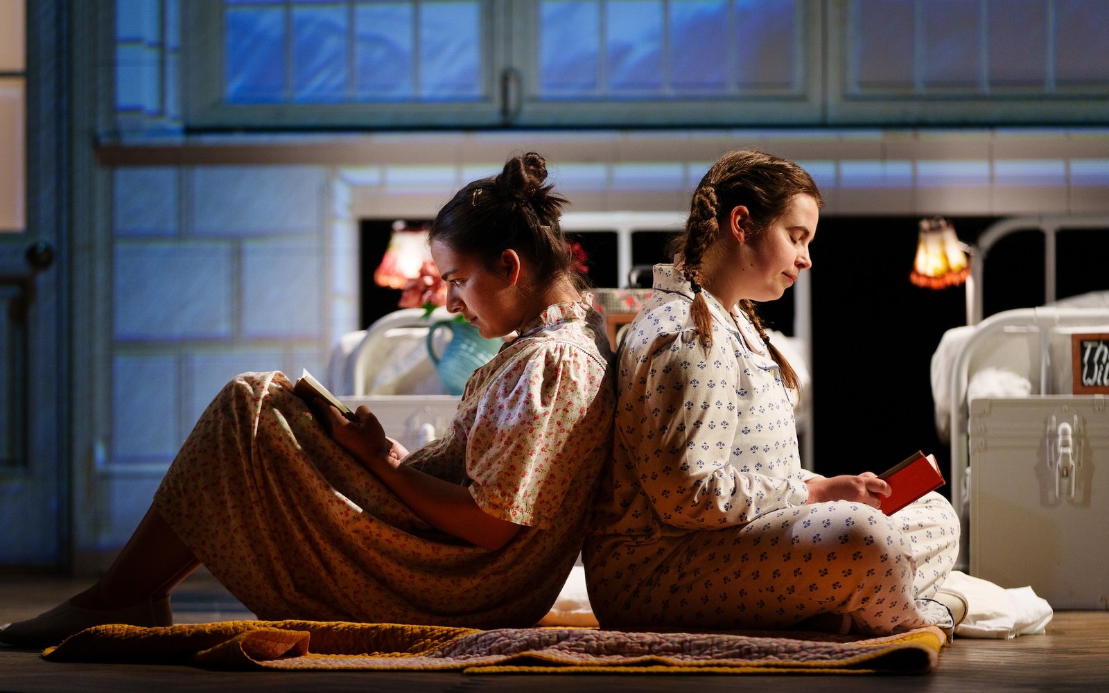Two girls sit back to back wearing nightdresses and reading books