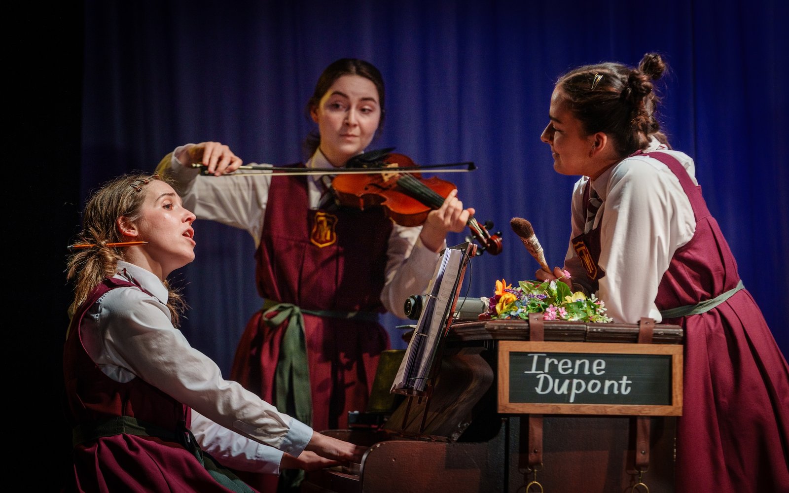 Three girls stand around a piano, one is playing the piano and singing and another is playing a violin