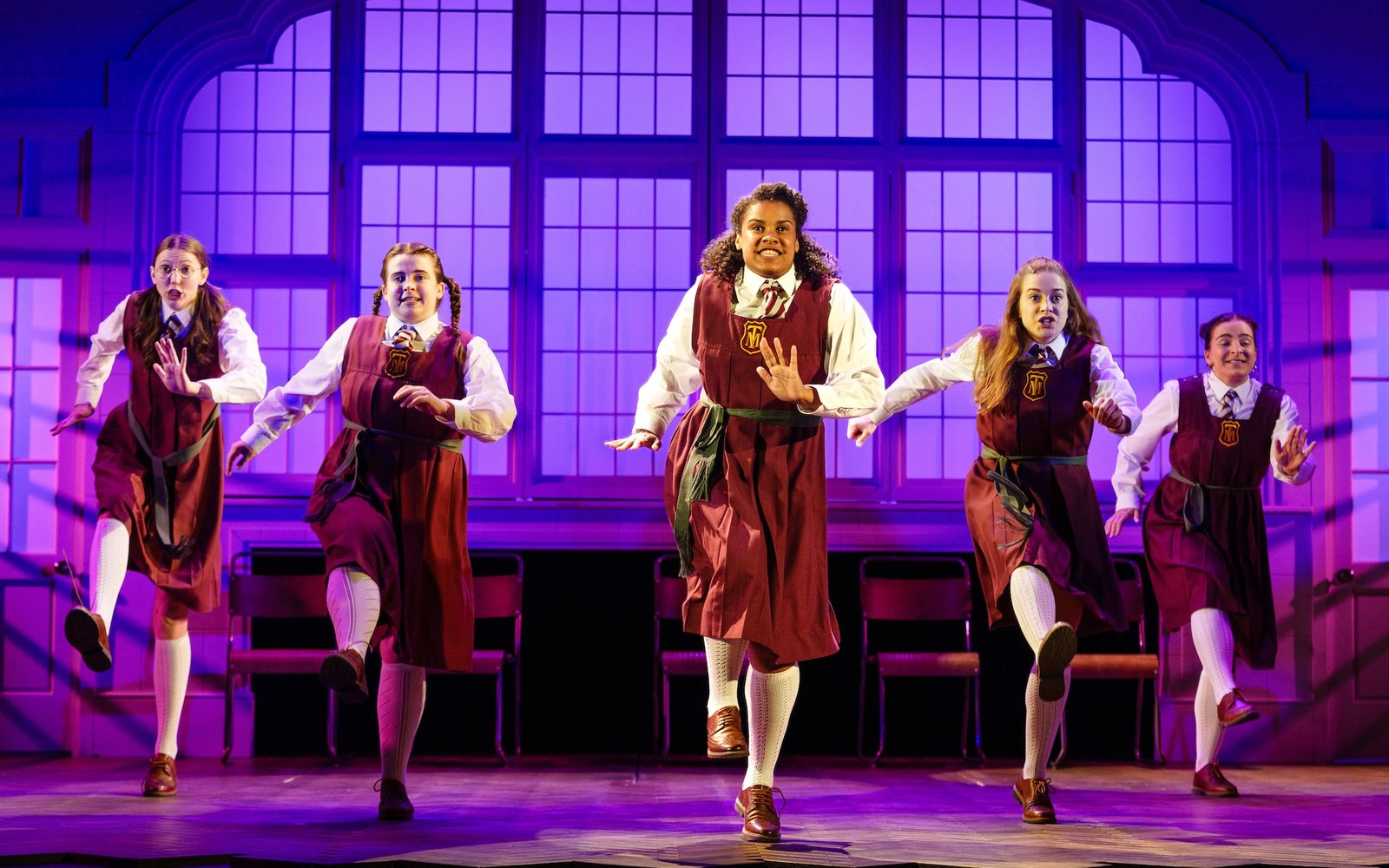 Five girls in traditional boarding school uniform dresses dance in unison in a classroom