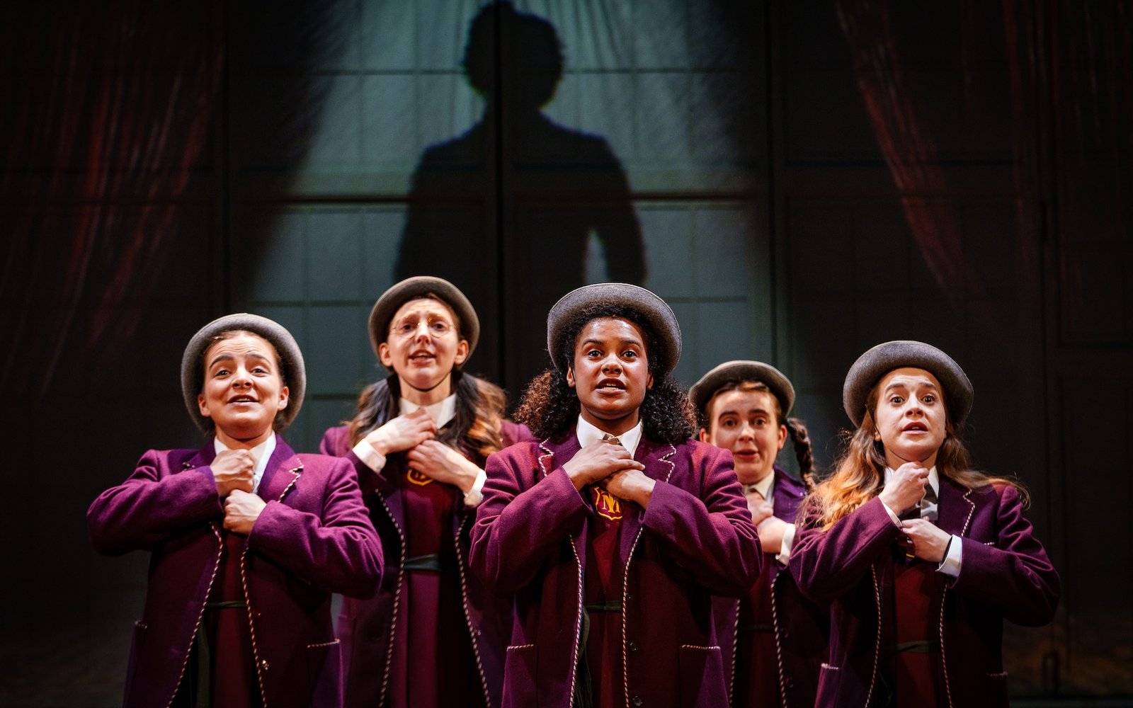 Five girls stand in traditional school uniforms and boater hats, there is a silhouette of a person behind them