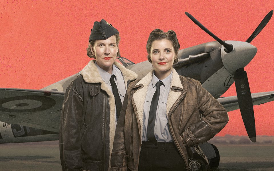 Two women in World War 2 bomber jackets stand in front of a propeller plane