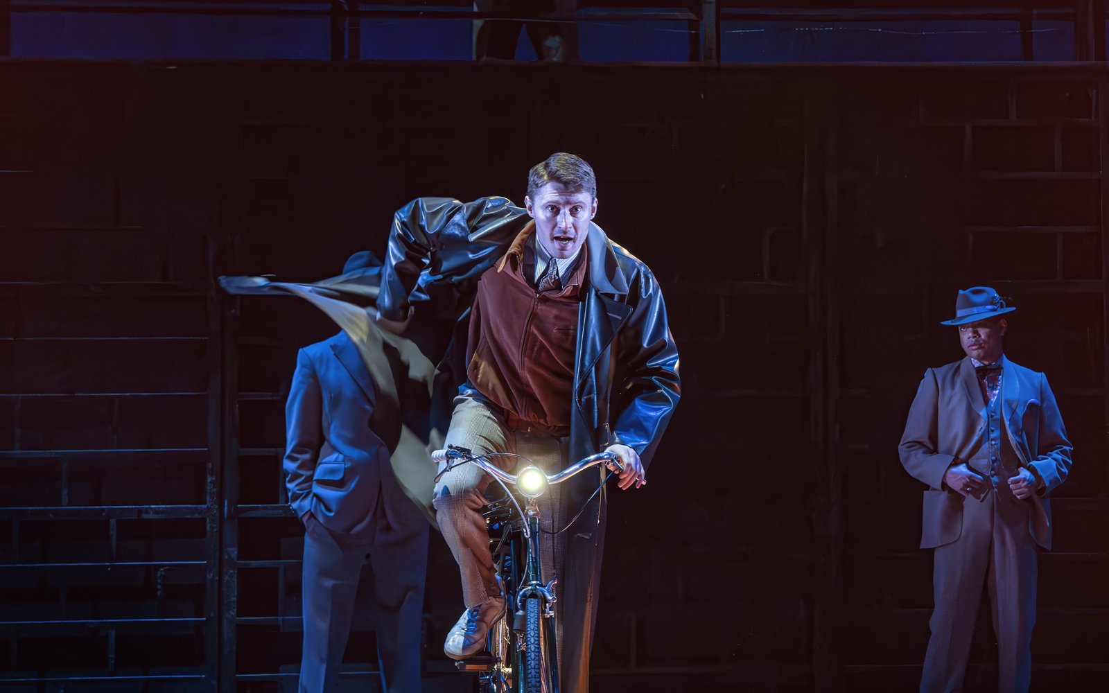 A man in a trench coat rides a bicycle while shadowy figures watch from behind him.
