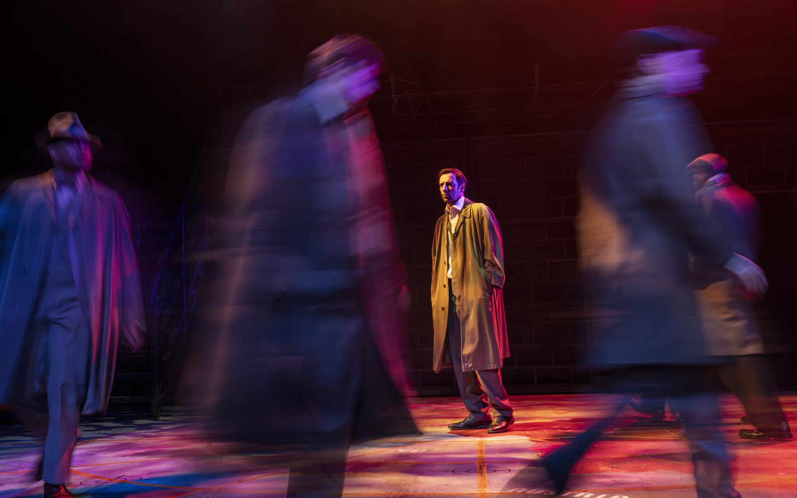 A solitary man in a coat stands still in the centre as blurred figures rush past him.