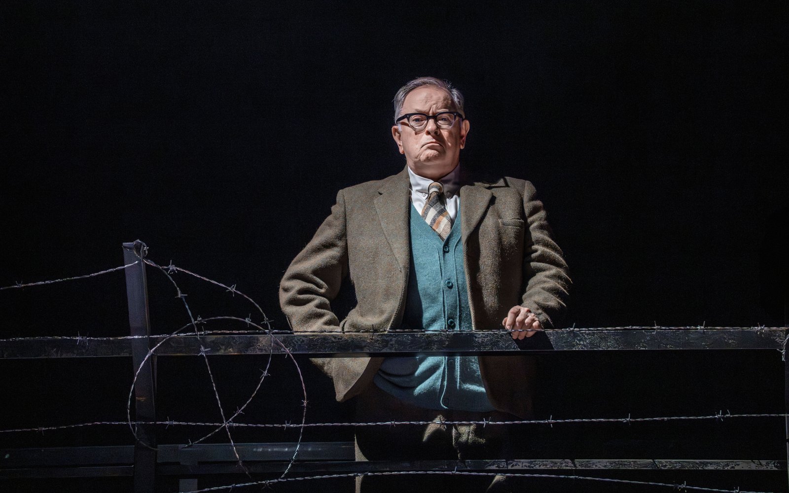 A man in glasses and a blazer looks out disapprovingly from behind a railing.