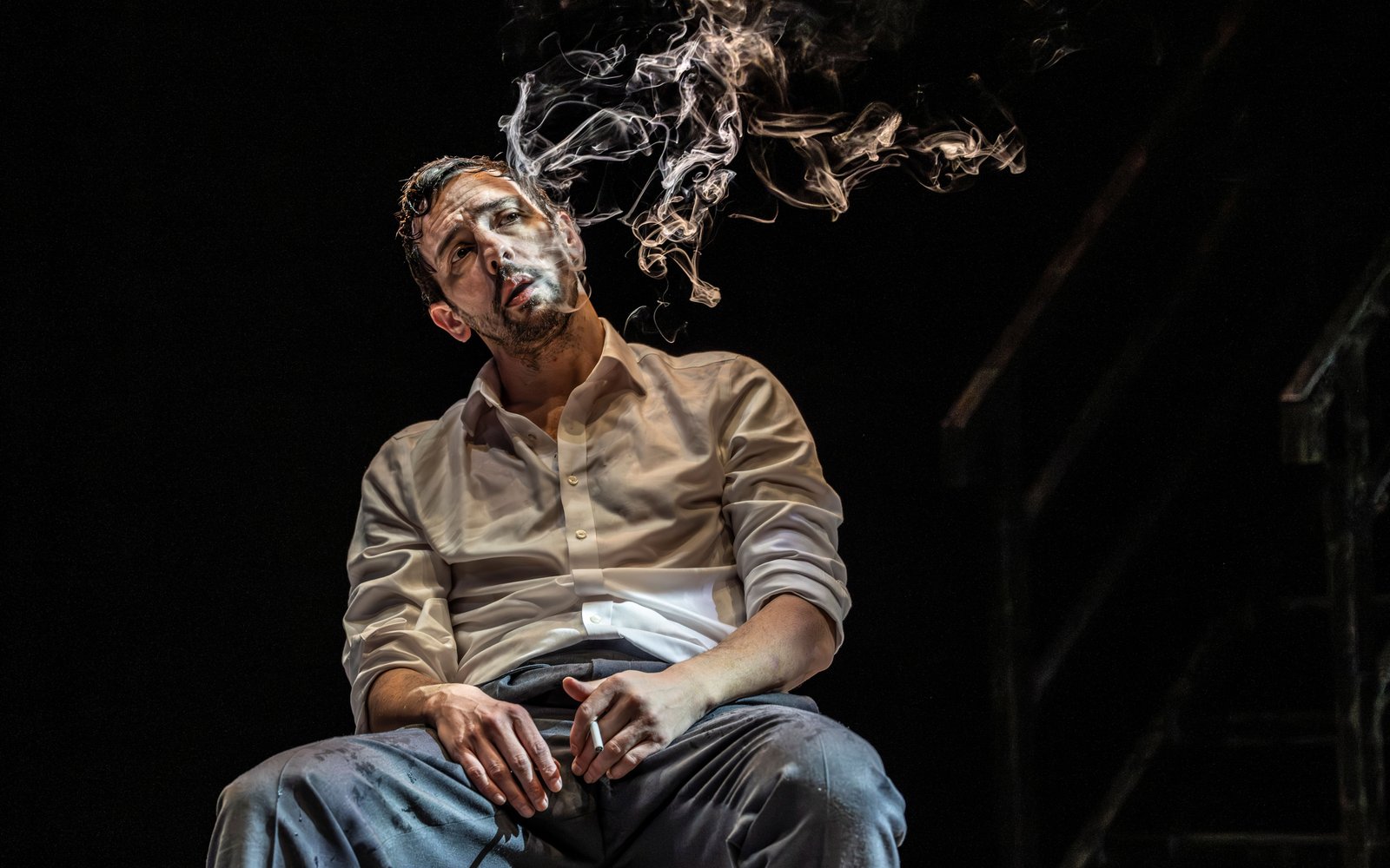 A man slouches in a chair holding a cigarette, with smoke in the air around him.