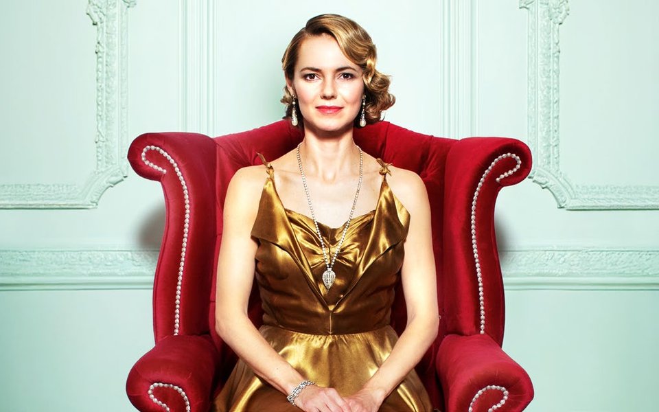 A glamorous woman in a gold dress sits on a grand red armchair