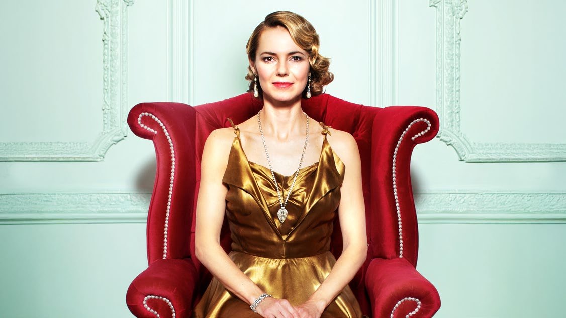 A glamorous woman in a gold dress sits on a grand red armchair