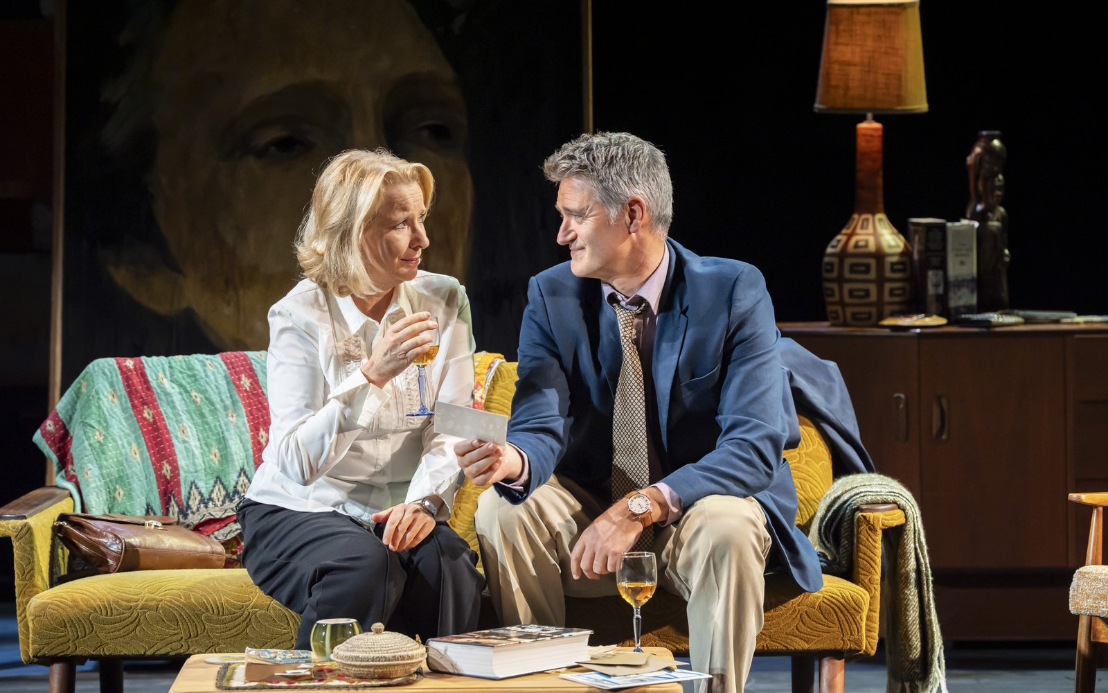 A man and a woman sit together on a sofa drinking wine, the man is holding a photograph
