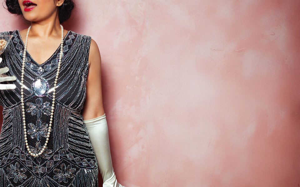 A glamorous 1920s flapper girl wearing pearls and sequins stands against a pink backdrop