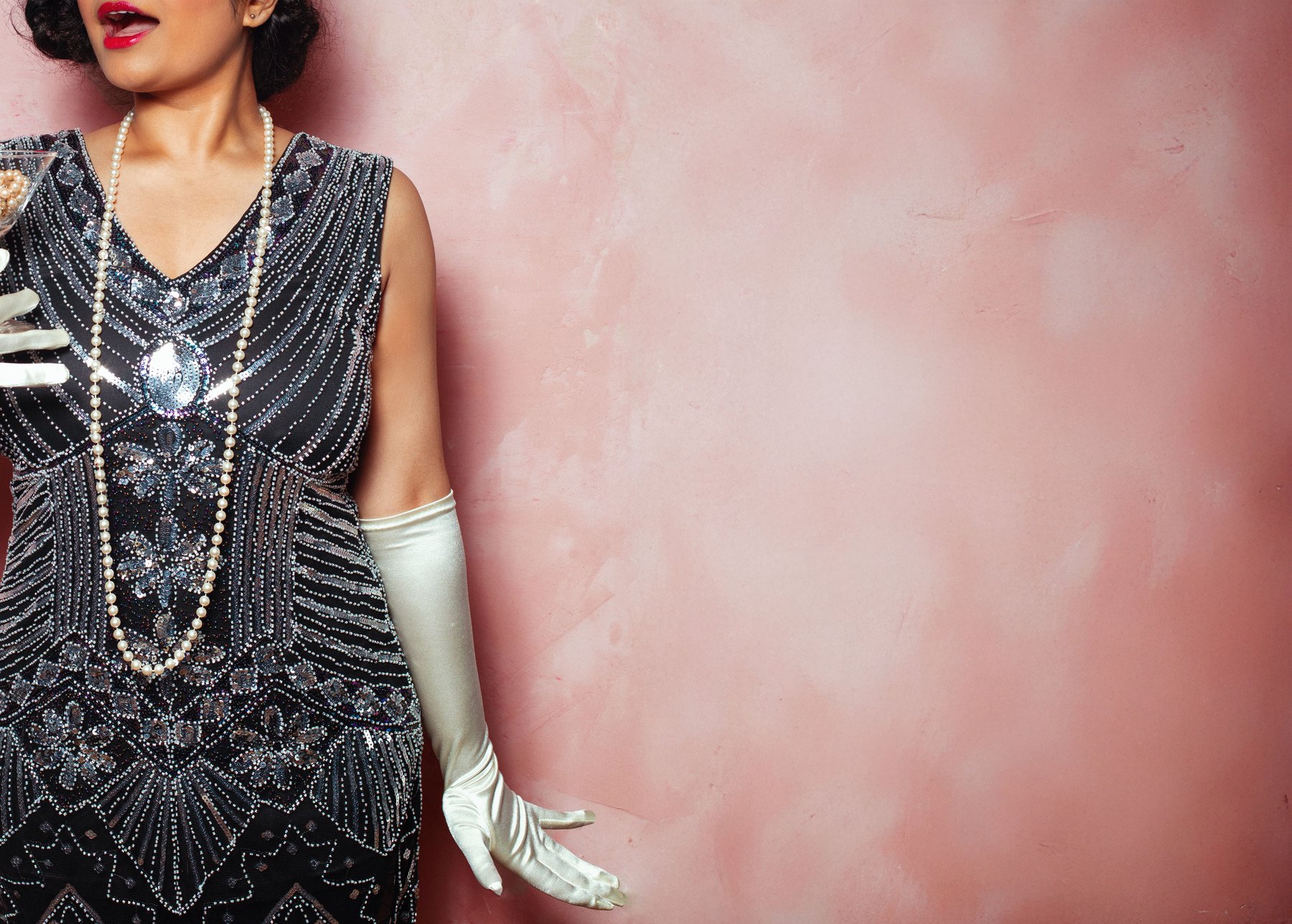 A glamorous 1920s flapper girl wearing pearls and sequins stands against a pink backdrop