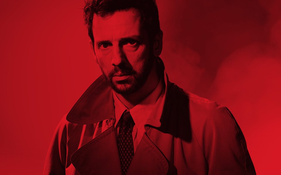 A man wearing a suit and jacket stares intensely, the image is washed with red