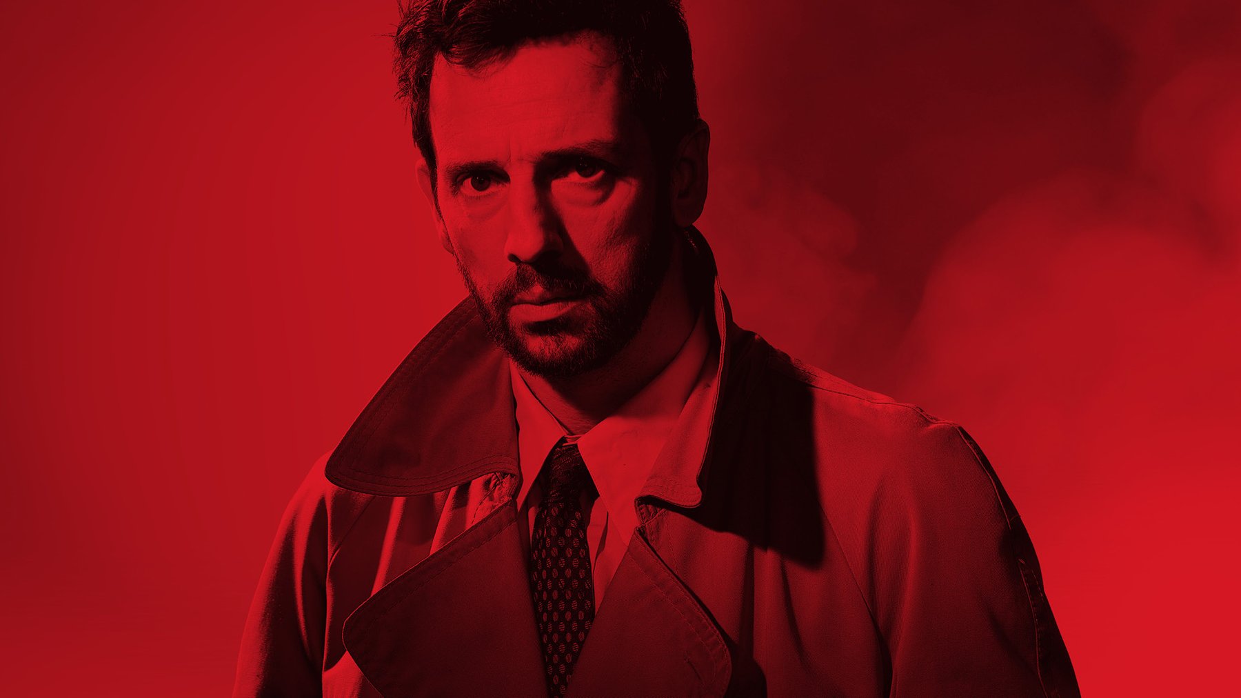 A man wearing a suit and jacket stares intensely, the image is washed with red