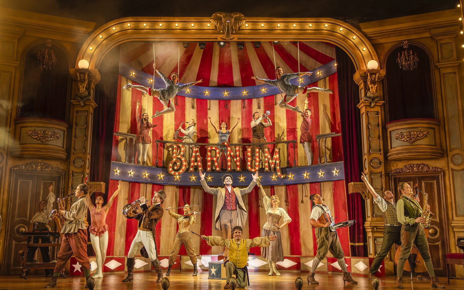A circus troupe performing on a brightly lit stage, people are playing instruments, hanging in trapeze and smiling with their arms outstretched