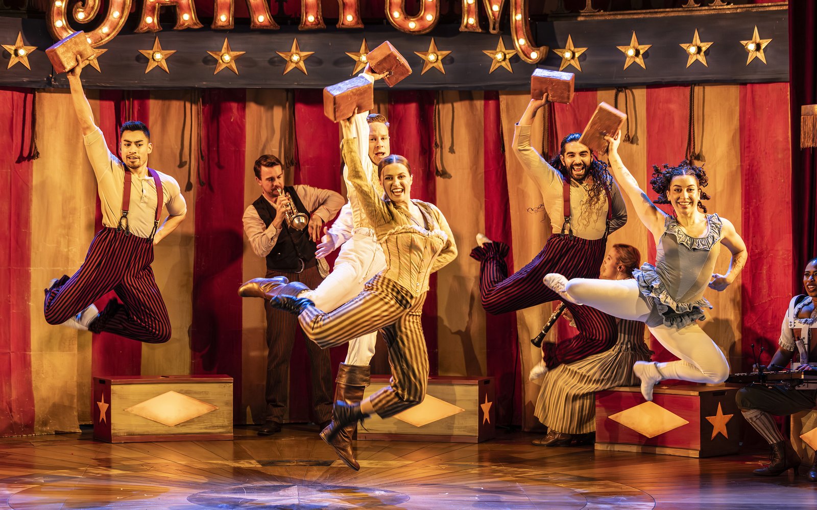 A group of circus performers dance in unison, they are wearing striped dungarees circus clothes and people are playing instruments behind them