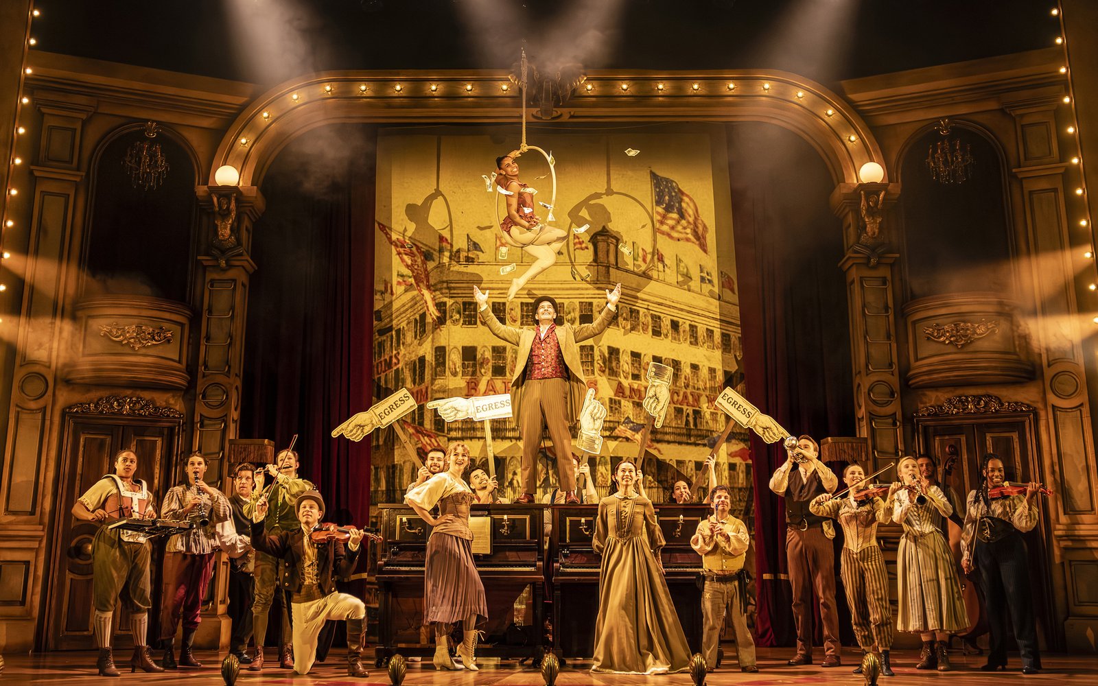 A large group of people stand smiling together on stage playing instruments, a man stands on top of a grand piano and above him a woman hangs in a trapeze hoop