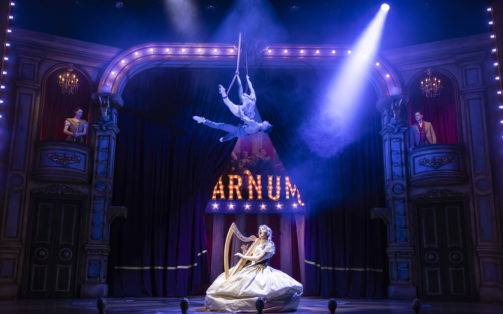 A woman plays a harp and sings passionately on stage, above her two people hang suspended in trapeze