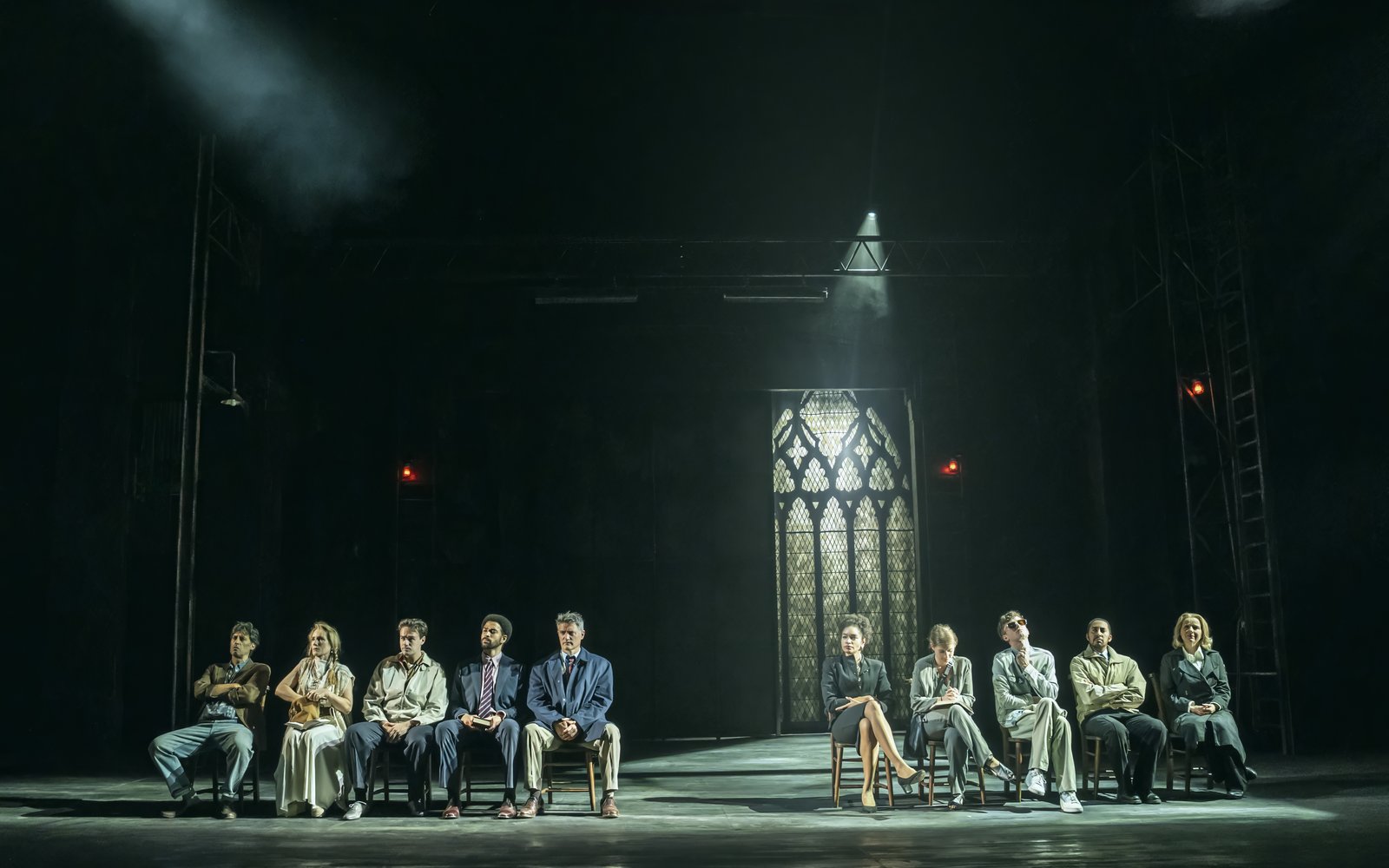 A group of men and woman sit on chairs in a church, there is a large stained-glass window behind them