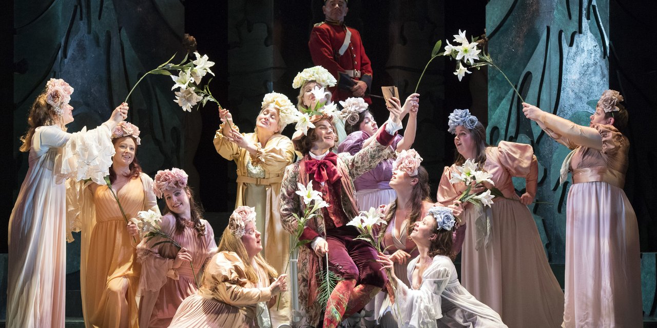 A group of women surround a man in regal costume, they waft flowers over him