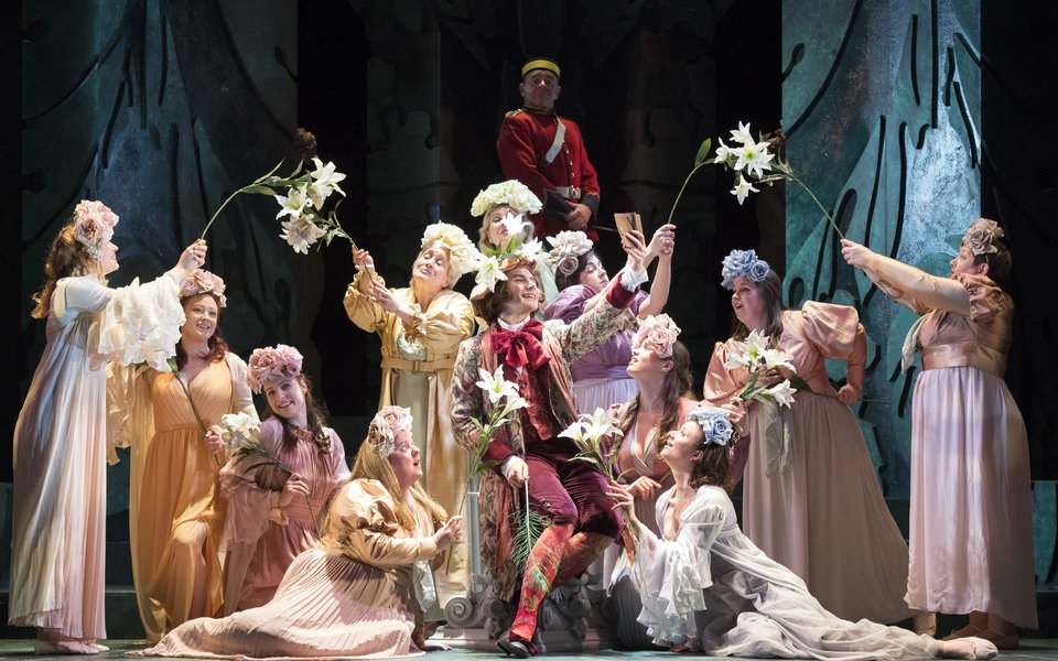 A group of women surround a man in regal costume, they waft flowers over him