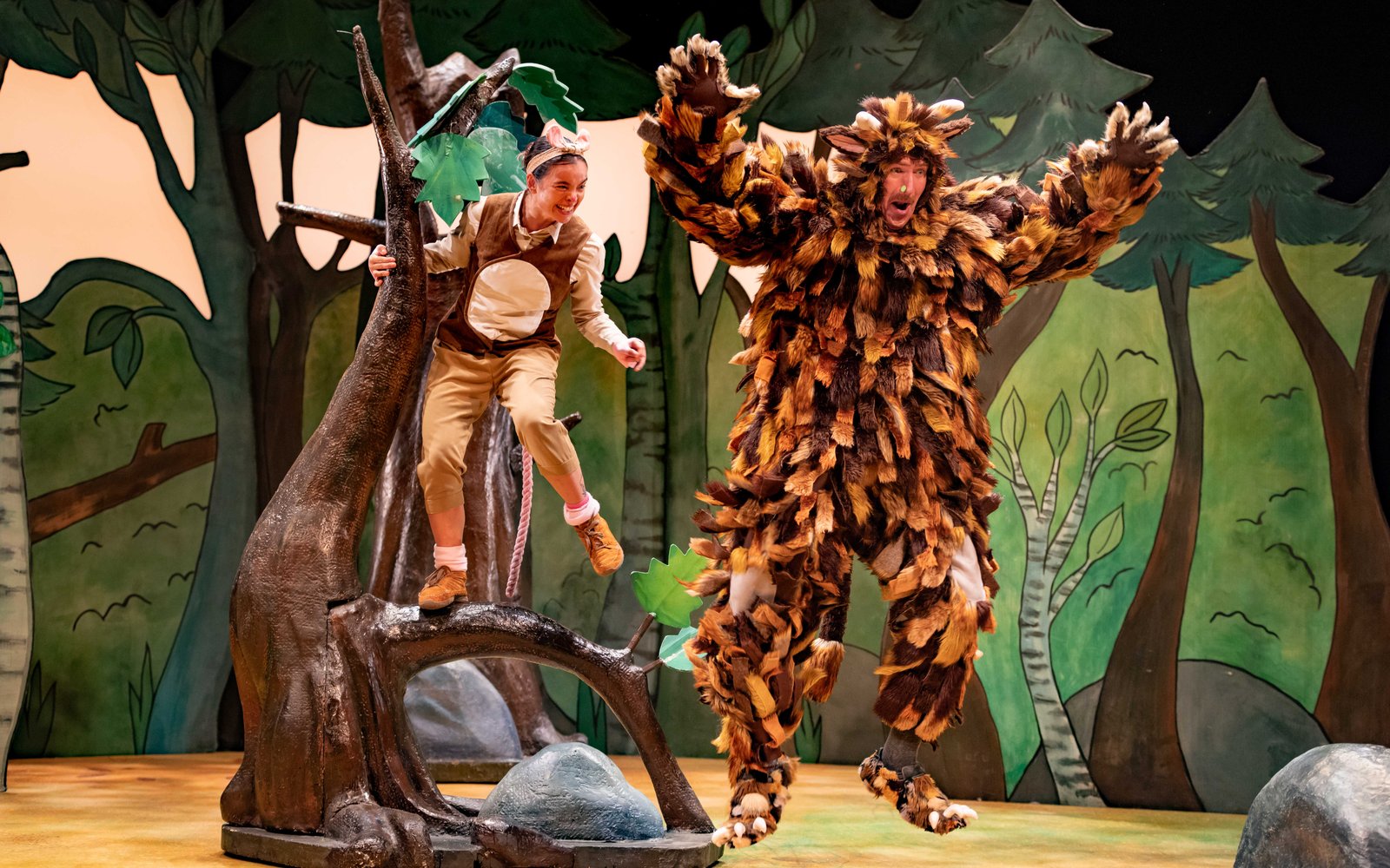 A man in a big fluffy brown Gruffalo costume jumps into the air in shock, a woman with mouse ears on is behind him standing on a tree stump
