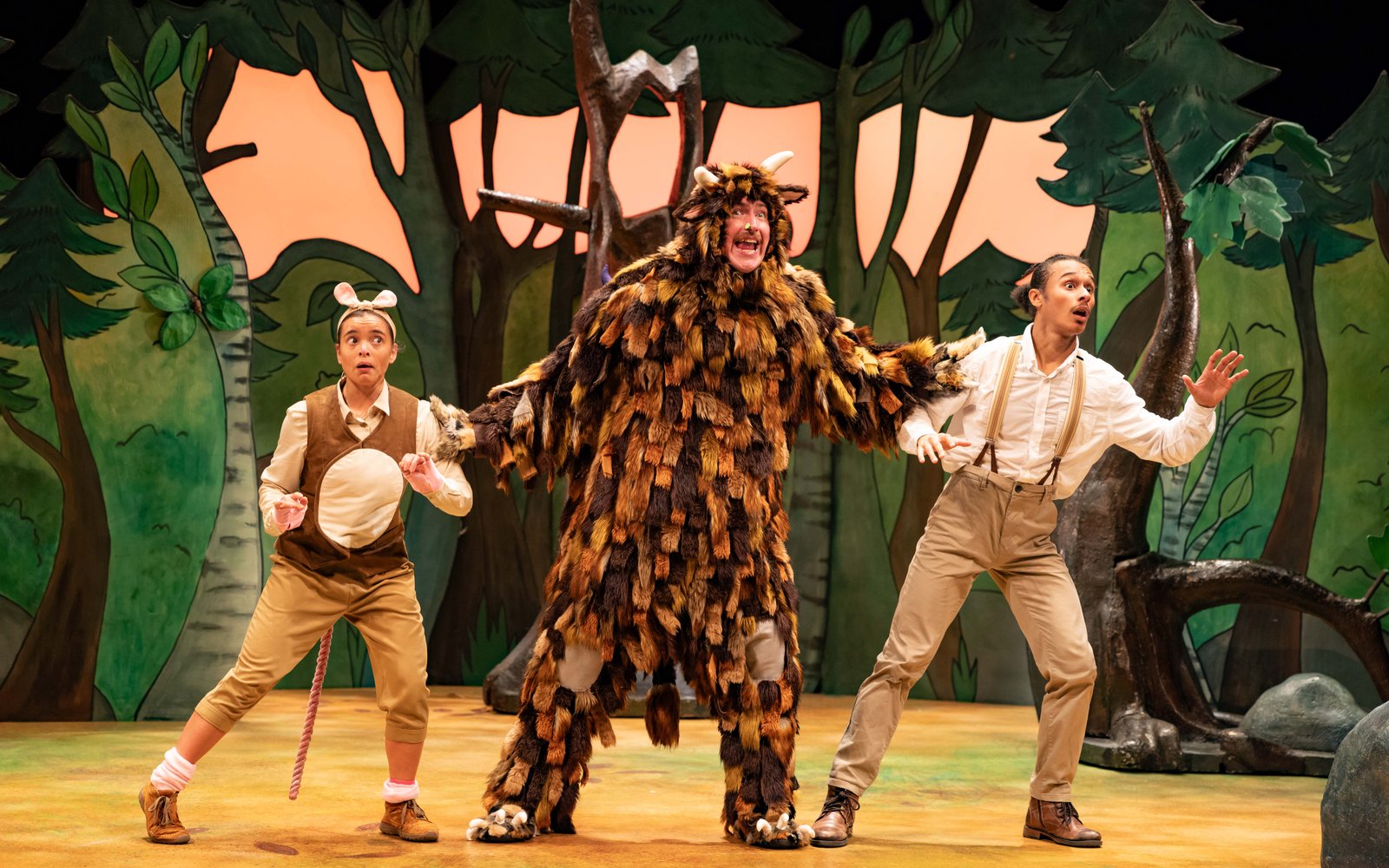 A man in a big fluffy brown Gruffalo costume stands between a man and a woman in beige trousers and shirts, they are in a forest