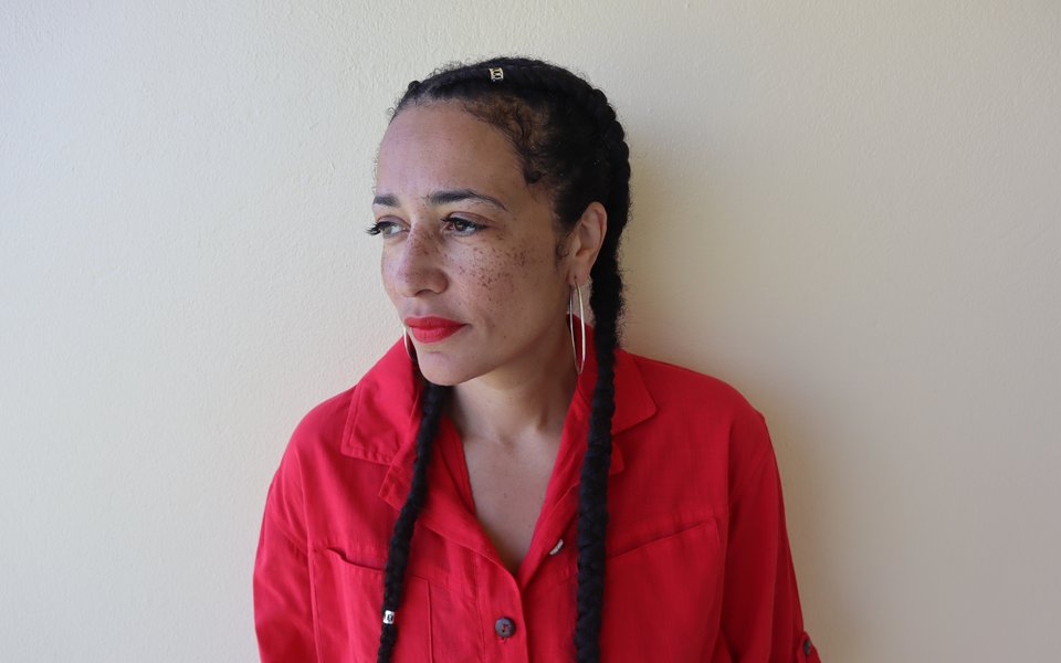 A woman stands against a white wall, her hair is in braids and she wears a red jumpsuit