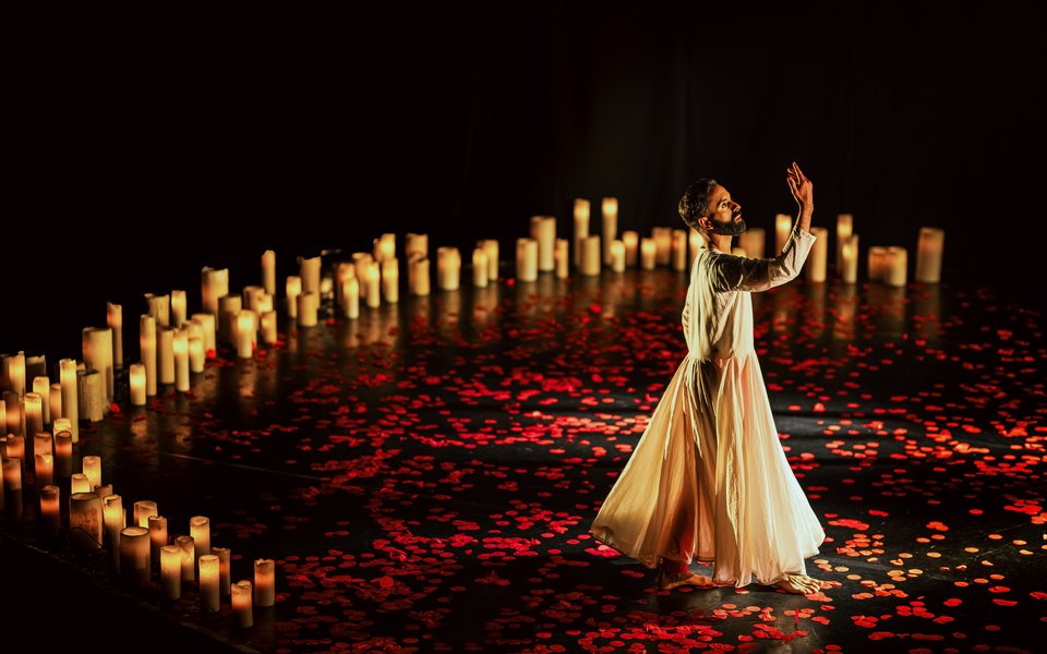 A dancer stands in a dark space wearing a white gown, red petals are on the floor and candles are lit behind him