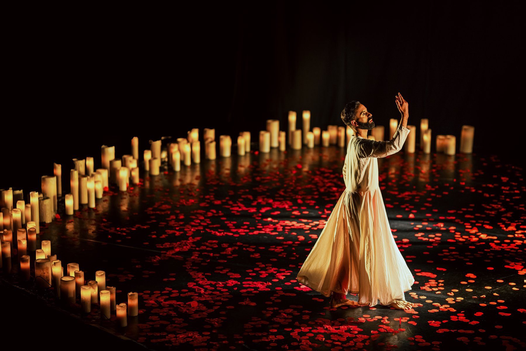 A dancer stands in a dark space wearing a white gown, red petals are on the floor and candles are lit behind him